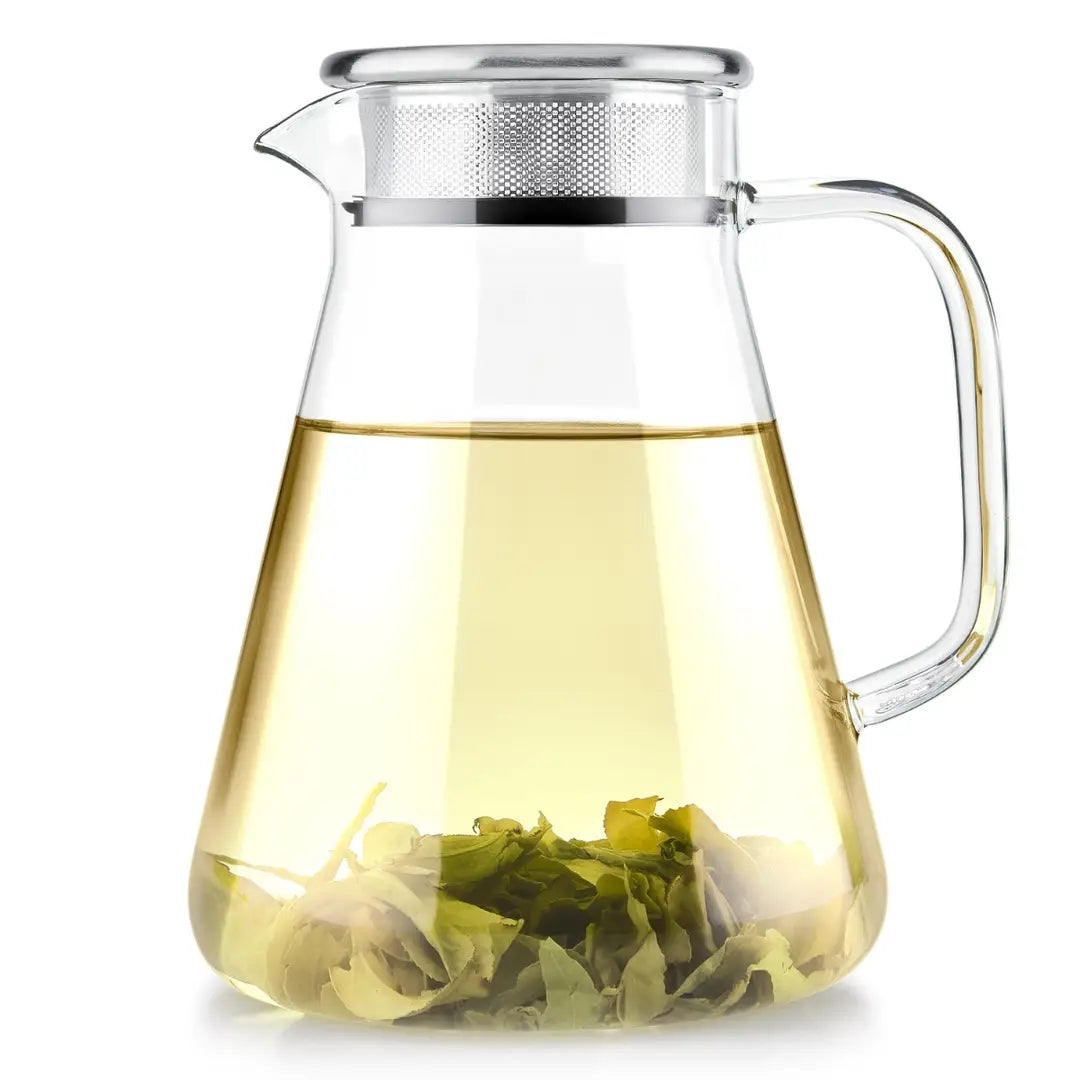 Clear glass teapot pitcher with a metal strainer lid, filled with pale yellow tea and green tea leaves at the bottom, on a white background.