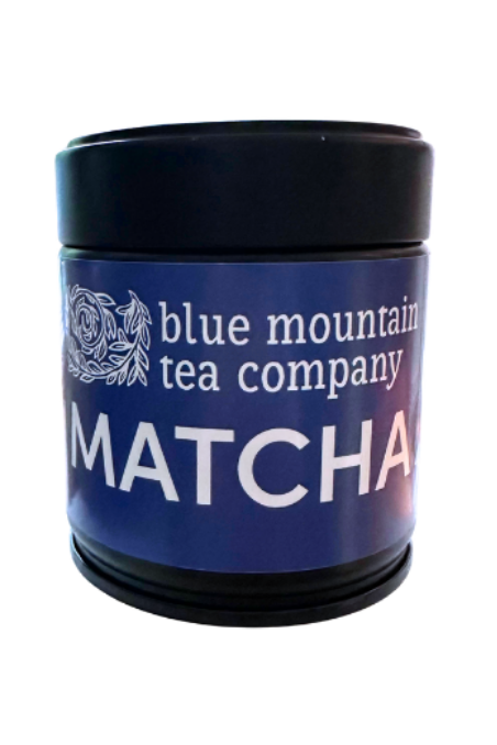 A black cylindrical tin of Blue Mountain Tea Company Matcha on a plain white background.